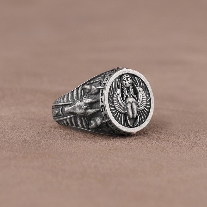 May include: Silver ring with an intricate design featuring an Egyptian scarab beetle with wings and the Eye of Horus. The ring band is decorated with hieroglyphs.