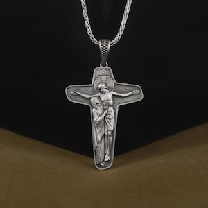 May include: A silver pendant necklace featuring a detailed depiction of Jesus Christ on the cross with Mary, his mother, standing beside him. The pendant is attached to a silver chain.