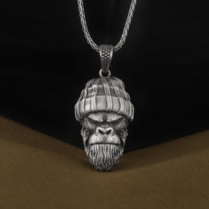 May include: Silver pendant necklace featuring a detailed gorilla head wearing a beanie hat. The chain is a silver rope chain.