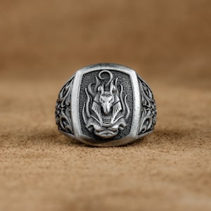 Puede incluir: Anillo de plata con un diseño ornamentado que presenta la cabeza de un dios egipcio con una corona y llamas.