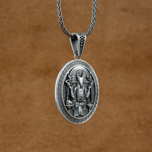 May include: Silver pendant necklace featuring a detailed depiction of a horned deity with outstretched arms holding a scale in each hand. The pendant is surrounded by a Greek key border.
