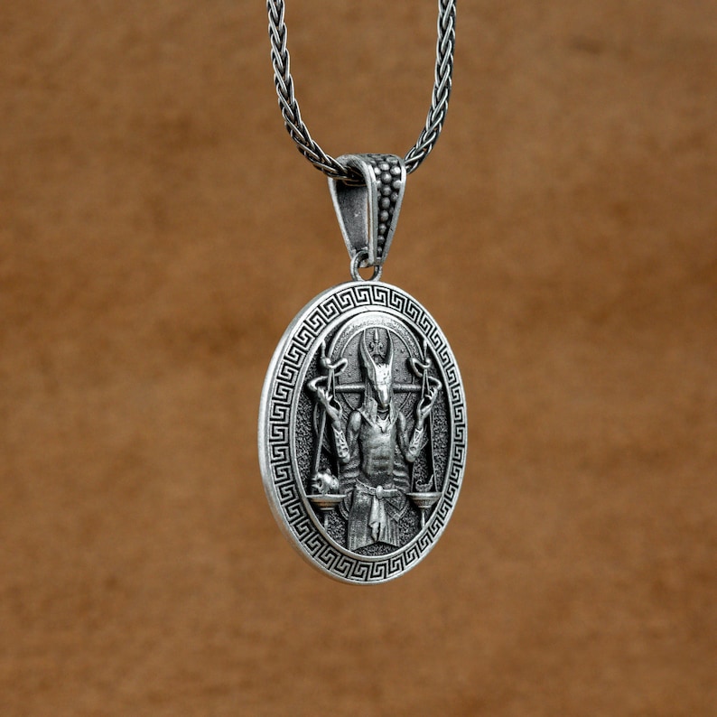 May include: Silver pendant necklace featuring a detailed depiction of a horned deity with outstretched arms holding a scale in each hand. The pendant is surrounded by a Greek key border.