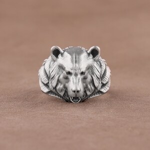 May include: A silver ring with a detailed bear head design. The bear's mouth is open, showing its teeth.