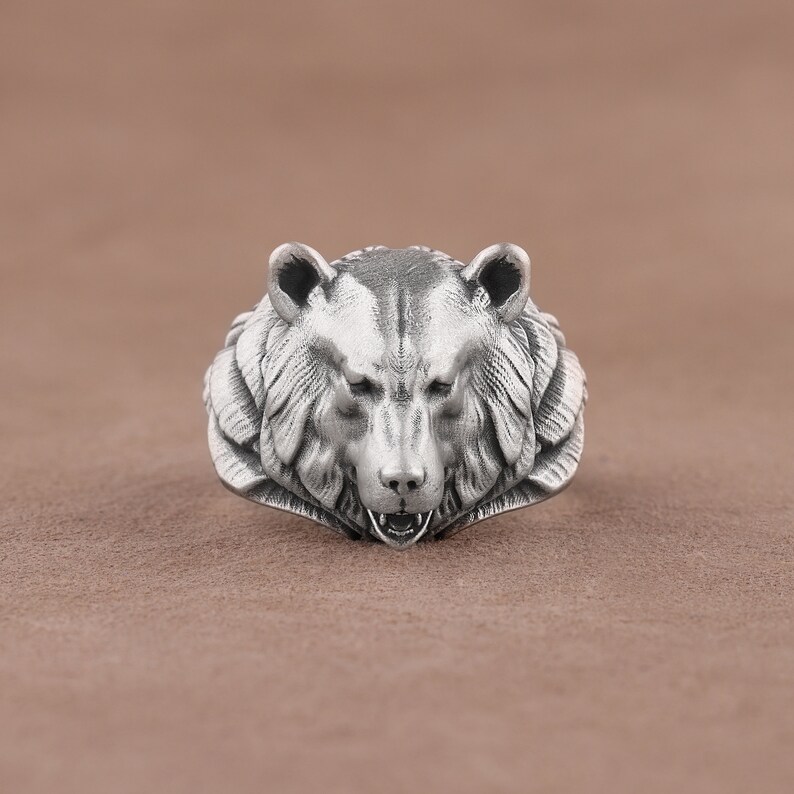 May include: A silver ring with a detailed bear head design. The bear's mouth is open, showing its teeth.