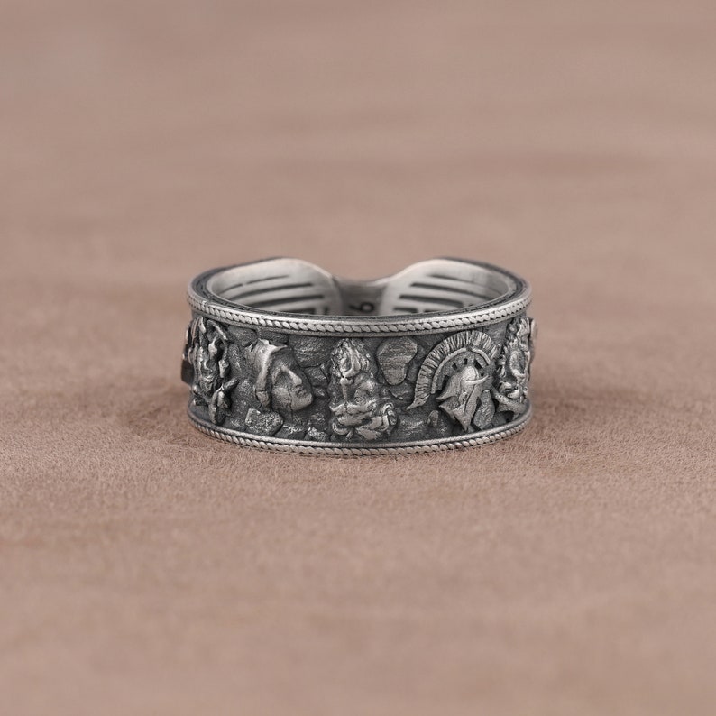 K&ouml;nnte beinhalten: Ein silberner Ring mit einem breiten Band, das mit einem komplizierten Design aus mehreren Gesichtern und einem Helm verziert ist. Der Ring liegt auf einem braunen Hintergrund.