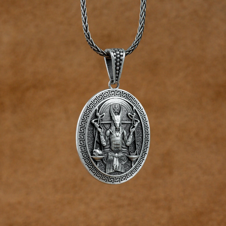 May include: Silver pendant necklace featuring an intricate design of a horned figure holding a scale. The pendant is oval shaped and has a Greek key border.