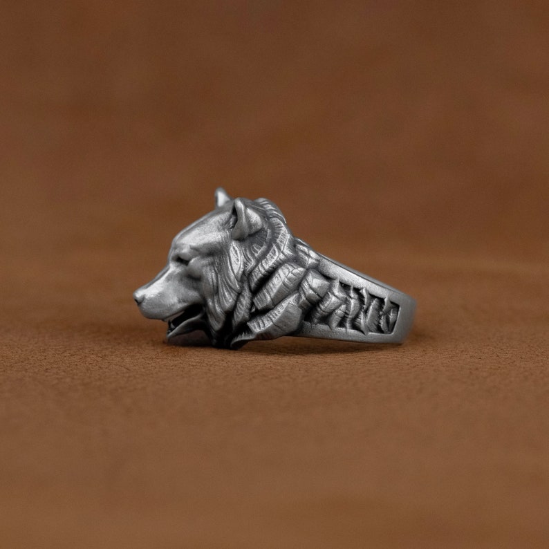 May include: A silver ring with a wolf head design. The wolf's head is detailed and realistic, with fur and teeth visible. The band of the ring is also decorated with a pattern.