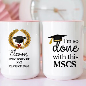 May include: Two white ceramic mugs with light pink handles and rims. One mug features a black graduation cap, a gold laurel wreath, a diploma, and the text Eleanor, UNIVERSITY OF XYZ, CLASS OF 2026. The other mug displays a graduation cap and the text I'm so done with this MSCS.
