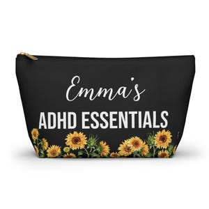 May include: A black zippered pouch featuring the white text "Emma's ADHD ESSENTIALS" printed in the center. The bottom portion of the pouch is decorated with a horizontal border of yellow sunflowers with green stems and leaves against the black background.