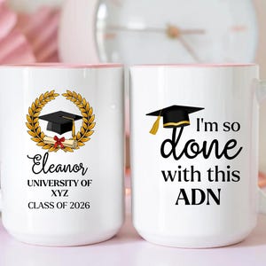 May include: Two white ceramic mugs with light pink handles and rims. One mug features a gold laurel wreath, a black graduation cap, a diploma, and the text Eleanor, UNIVERSITY OF XYZ, CLASS OF 2026. The other mug displays a black graduation cap and the text I'm so done with this ADN.