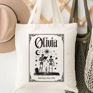 Puede incluir: Un bolso tote de color crema con un gráfico negro que presenta esqueletos, una luna y estrellas. La bolsa tiene el nombre "Olivia" y el texto "Book Lover Since 1988". Un sombrero color canela y dos bolsas más cuelgan de un perchero de madera.