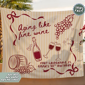 Puede incluir: Un tapiz a rayas beige y blanco con la frase "Aging like fine wine" en borgoña. El diseño incluye copas de vino, una botella, uvas y una barrica. El texto dice "Fort Lauderdale, FL Emma's 30th Birthday".