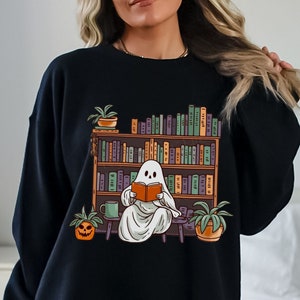 Puede incluir: Sudadera negra con un gráfico de un fantasma leyendo un libro en un entorno de biblioteca. El fantasma es blanco y las estanterías de la biblioteca están llenas de libros coloridos.