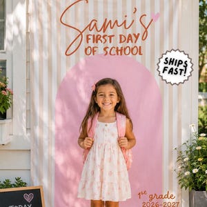 May include: A vertical fabric banner featuring beige and white vertical stripes with a large pink arch graphic. The text reads Sami's FIRST DAY OF SCHOOL and 1st grade 2026-2027. A small chalkboard sign nearby displays the text TODAY IS A GOOD DAY TO LEARN.