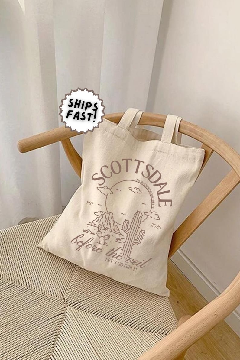 May include: Beige tote bag with the word "Scottsdale" in a brown font, along with a desert landscape graphic. The text "before the veil" and "Let's go girls!" are also printed on the bag. The bag is sitting on a wooden chair.