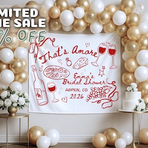May include: A bridal shower backdrop with a white banner featuring red illustrations of wine, food, and text that reads "That's Amore" and "Emma's Bridal Shower, Aspen, CO 2026." The scene is decorated with gold and white balloons, a cake, and floral arrangements.