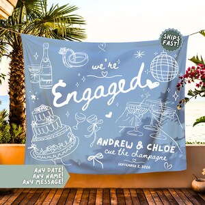 May include: A light blue fabric backdrop featuring white line art illustrations of a champagne bottle, wedding cake, disco ball, and rings. The text reads "we're Engaged", "ANDREW & CHLOE", "cue the champagne", and "SEPTEMBER 2, 2026" with small heart accents.