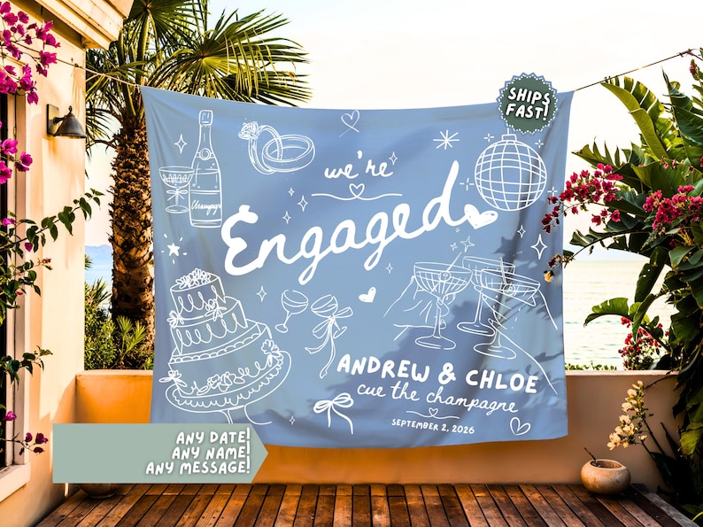 May include: A light blue fabric backdrop featuring white line art illustrations of a champagne bottle, wedding cake, disco ball, and rings. The text reads "we're Engaged", "ANDREW & CHLOE", "cue the champagne", and "SEPTEMBER 2, 2026" with small heart accents.