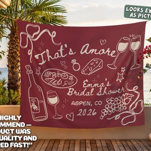 May include: Maroon banner with white illustrations of wine bottles, glasses, and food, with the text "That's Amore" and "Emma's Bridal Shower, Aspen, CO 2026". The banner is hanging outdoors.