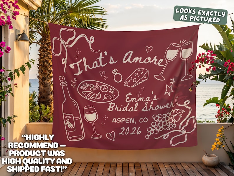 May include: Maroon banner with white illustrations of wine bottles, glasses, and food, with the text "That's Amore" and "Emma's Bridal Shower, Aspen, CO 2026". The banner is hanging outdoors.