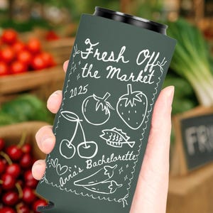 May include: Dark green can cooler with white text and illustrations. The text reads "Fresh Off the Market 2025" with drawings of a cherry, tomato, strawberry, fish, and pepper. The text "Anna's Bachelorette" is also visible.