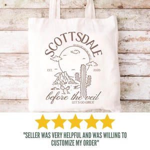 May include: Off-white tote bag with a brown graphic design featuring the word "Scottsdale" in a circular design, a cactus, and the text "before the veil". The bag has two handles and is made of a canvas-like material.