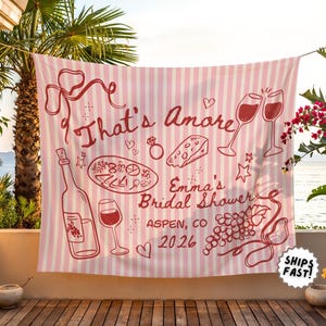 May include: A pink and white striped banner with the words "That's Amore" and "Emma's Bridal Shower Aspen, CO 2026" in red. The banner features illustrations of wine, food, and celebratory elements.