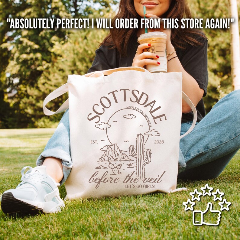 May include: A cream-colored tote bag with the word "SCOTTSDALE" in a circular design, featuring a desert landscape illustration. The text "before the veil" and "LET'S GO GIRLS!" are also visible. The bag is held by a person.
