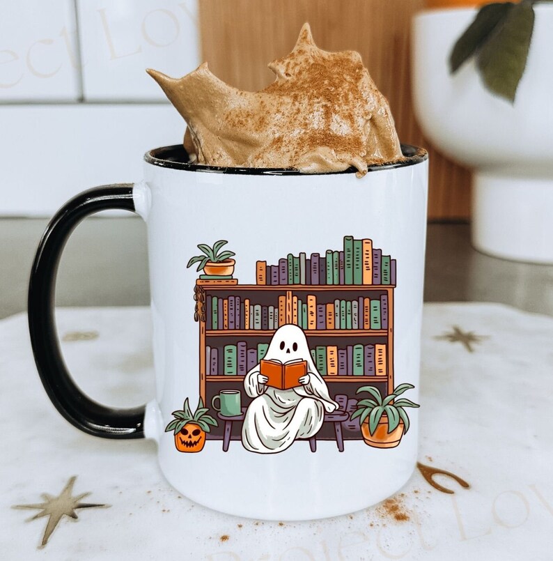 white 11oz ceramic mug with black inside and black handle, this ghost mug design has a cottagecore ghost reading a book with bookshelf and coffee mug, perfect gift for book lovers and a great ghost mug for a gothic friend.