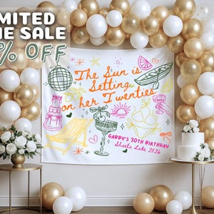 May include: A party backdrop with gold and white balloons, a white banner with colorful illustrations, and a two-tiered cake. The banner reads "The Sun is Setting on her Twenties" and "Gabby's 30th Birthday Shasta Lake 2026."