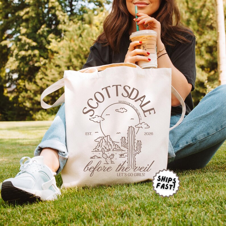 May include: Cream-colored tote bag with the word "SCOTTSDALE" in a circular design, featuring a desert landscape illustration. The text "before the veil" and "LET'S GO GIRLS!" are also visible. The bag has white handles and is held by a person.