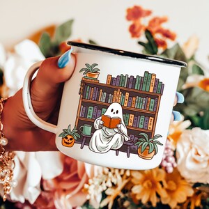 white camp enamel mug with black rim, this ghost mug design has a cottagecore ghost reading a book with bookshelf and coffee mug, perfect gift for book lovers and a great ghost mug for a gothic friend.