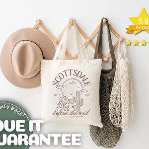 May include: A beige fedora hat, a white tote bag with "SCOTTSDALE before the veil" text, and two mesh bags hang on a wooden rack. The tote bag features a desert landscape design. A 5.0 rating star graphic is visible.