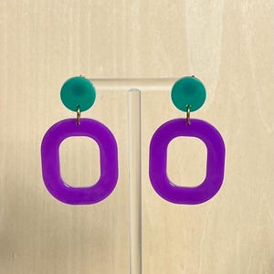 以下が含まれることがあります： 小さなティール色の円形スタッドと、角が丸い大きな紫色の長方形のフープが連結されたドロップピアス。各パーツは小さな金色の金属リングで繋がっており、幾何学的なデザインを形成しています。