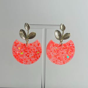 Può includere: Un paio di orecchini con un pendente a semicerchio riempito di glitter arancione neon brillante e macchioline iridescenti. Ogni pendente è fissato a un elemento in metallo dorato a forma di piccolo stelo con tre foglie sulla parte superiore.