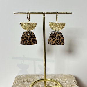 Puede incluir: Un par de pendientes dorados con una parte superior texturizada en forma de semicírculo y una parte inferior con estampado de leopardo. Los pendientes se exhiben en un soporte dorado para pendientes con una base circular. El fondo es de color neutro.