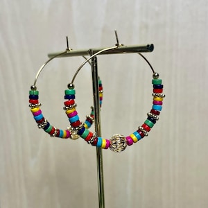 Op de afbeelding: Gouden oorringen versierd met kleurrijke kralen in rood, blauw, geel, groen en roze. De oorbellen hebben gouden accenten en een decoratieve gouden bol. De oorbellen worden op een gouden standaard getoond.