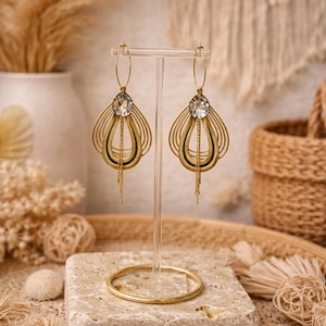 Op de afbeelding: Goudkleurige statement oorbellen met een druppelvormig ontwerp, met een grote, heldere kristal aan de bovenkant en delicate kettingfranjes. De oorbellen worden gepresenteerd op een transparante standaard, die hun ingewikkelde details en elegante stijl benadrukt.