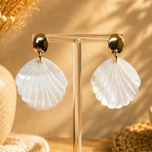 Peut inclure: Une paire de boucles d'oreilles pendantes composée d'un clou circulaire doré poli sur le dessus, relié à un grand pendentif en forme de coquillage blanc en éventail. Les coquillages ont une finition irisée lisse avec des stries verticales.