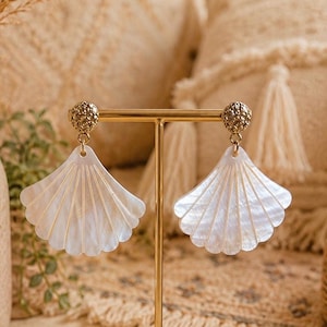 Peut inclure: Une paire de boucles d'oreilles élégantes en forme de coquillage blanc avec des accents dorés. Les boucles d'oreilles présentent un design détaillé en forme d'éventail avec des lignes verticales et un clou doré texturé en haut.