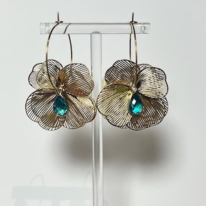 Op de afbeelding: Een paar goudkleurige draadbloemoorbellen. Elke oorbel heeft een bloemvorm met een turquoise druppelvormige edelsteen in het midden. De oorbellen hangen aan goudkleurige hoepels.