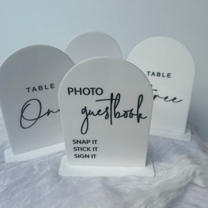 May include: Three white acrylic table signs with black calligraphy. The signs read "Table One", "Photo Guestbook", and "Table Three". The photo guestbook sign also includes the text "Snap it, Stick it, Sign it".