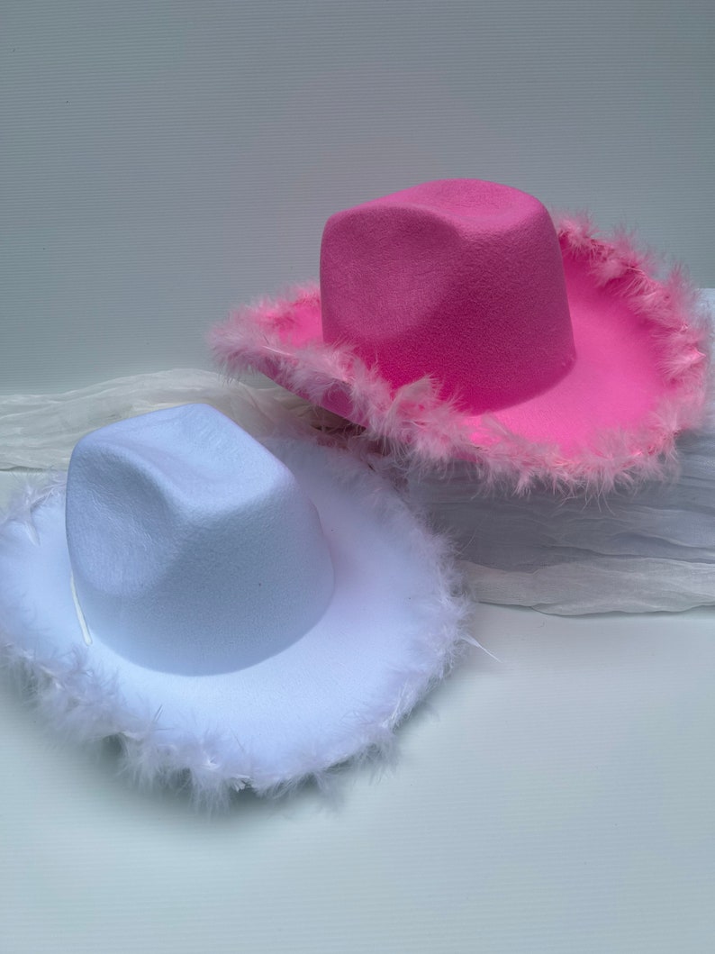 May include: Two cowboy hats with feather trim. One hat is white and the other is pink.
