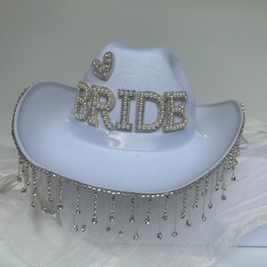 May include: White cowboy hat with a rhinestone fringe and the word "BRIDE" spelled out in rhinestones. A small rhinestone heart is on the top of the hat.