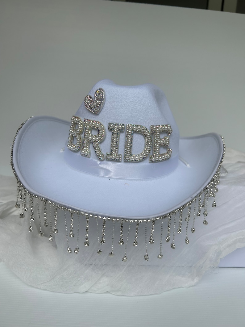 May include: White cowboy hat with a rhinestone fringe and the word "BRIDE" spelled out in rhinestones. A small rhinestone heart is on the top of the hat.