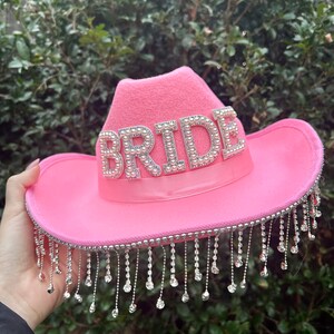 May include: Pink cowboy hat with the word "BRIDE" spelled out in rhinestones. The hat has a fringe of silver beads around the brim.