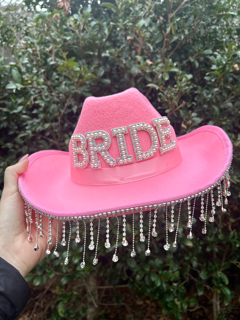 May include: Pink cowboy hat with the word "BRIDE" spelled out in rhinestones. The hat has a fringe of silver beads around the brim.
