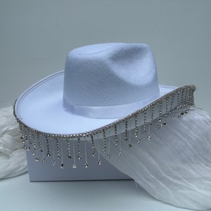 May include: A white cowboy hat with a silver rhinestone trim and fringe. The hat is sitting on a white surface.