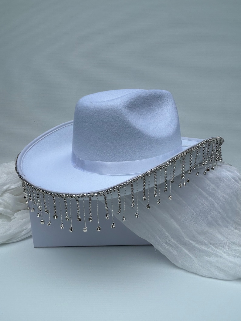 May include: A white cowboy hat with a silver rhinestone trim and fringe. The hat is sitting on a white surface.