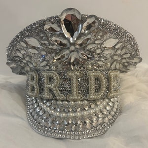 May include: A white bridal hat decorated with rhinestones and pearls. The hat has the word "BRIDE" spelled out in rhinestones and pearls.